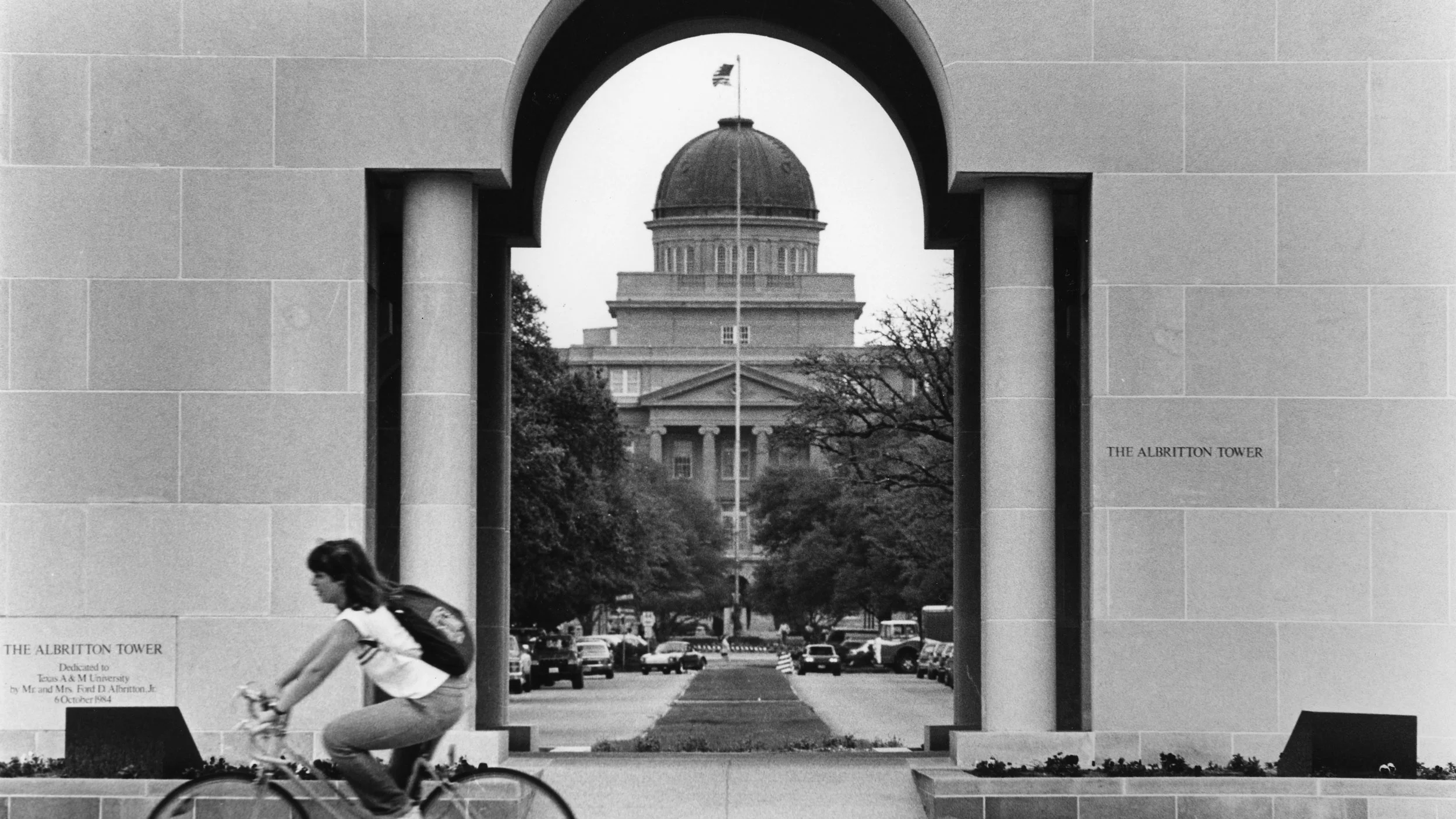 Black and white photo of cyclist passing through Albritton Tower archway, with Academic Building's domed tower visible in perfect frame beyond campus mall.