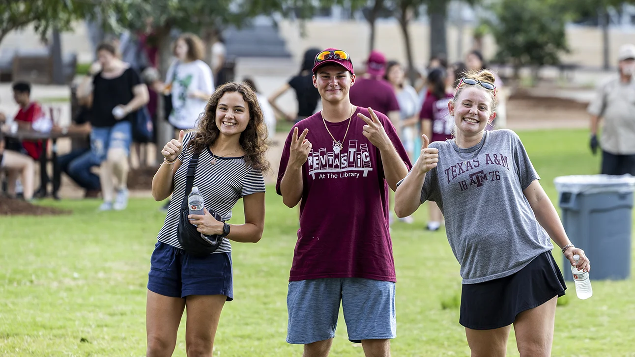 Three smiling Texas A&M students pose outdoors on campus, giving thumbs up and Aggie hand signs at a casual outdoor gathering.