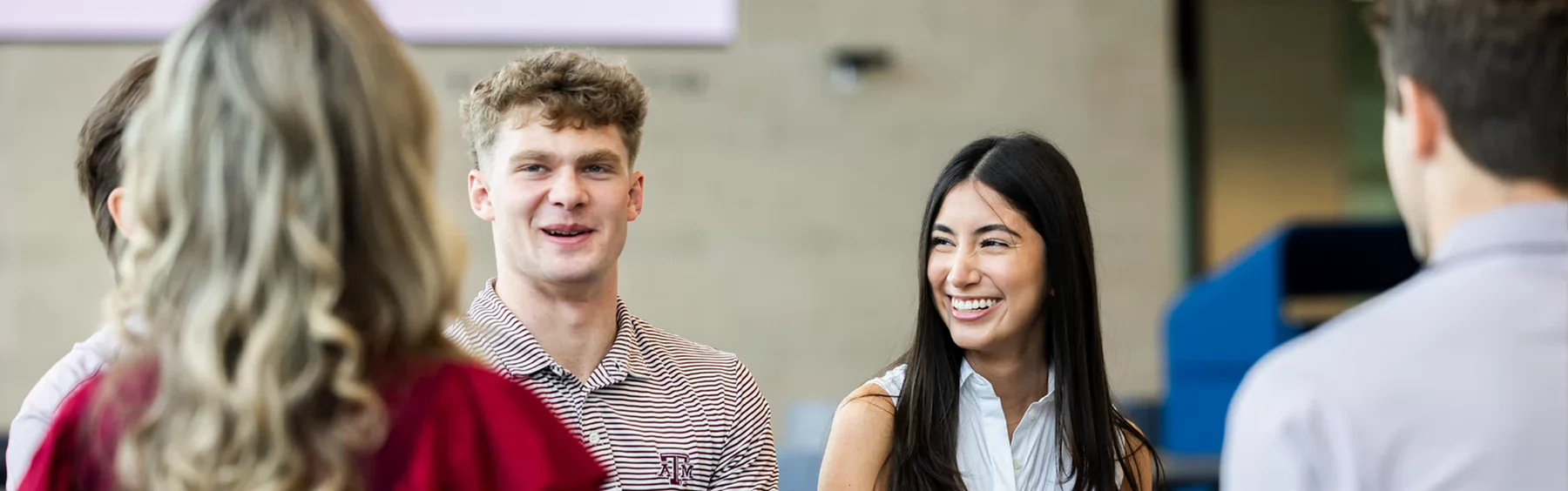 College students smile and converse in a campus building, with one wearing a Texas A&M striped polo shirt.
