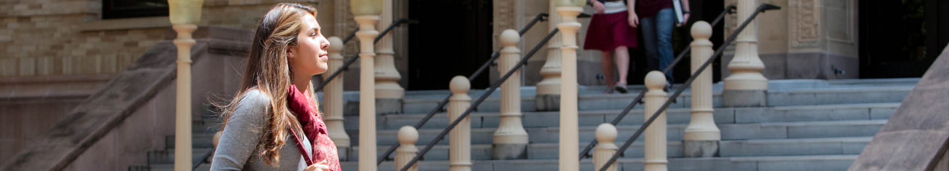 Young woman with long brown hair and pink scarf stands in profile before historic building with ornate stone columns and stairs visible in background.