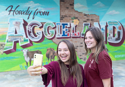 Two students take a photo in front of a mural that reads “Howdy from Aggieland.”