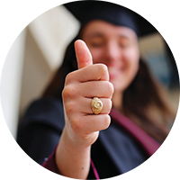 A student in a graduation gown makes a Gig'em thumbs up hand gesture.