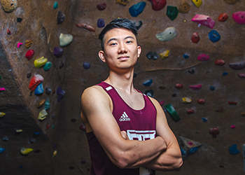 A student stands arms crossed in front of an indoor bouldering rock wall.