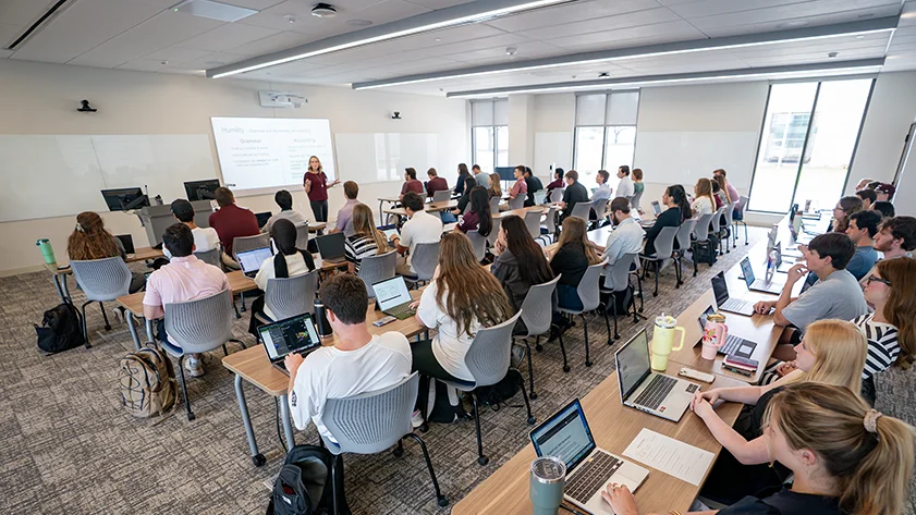 A instructor leads a large university classroom of students with laptops seated at modern tables, with a presentation displayed on a projector screen.